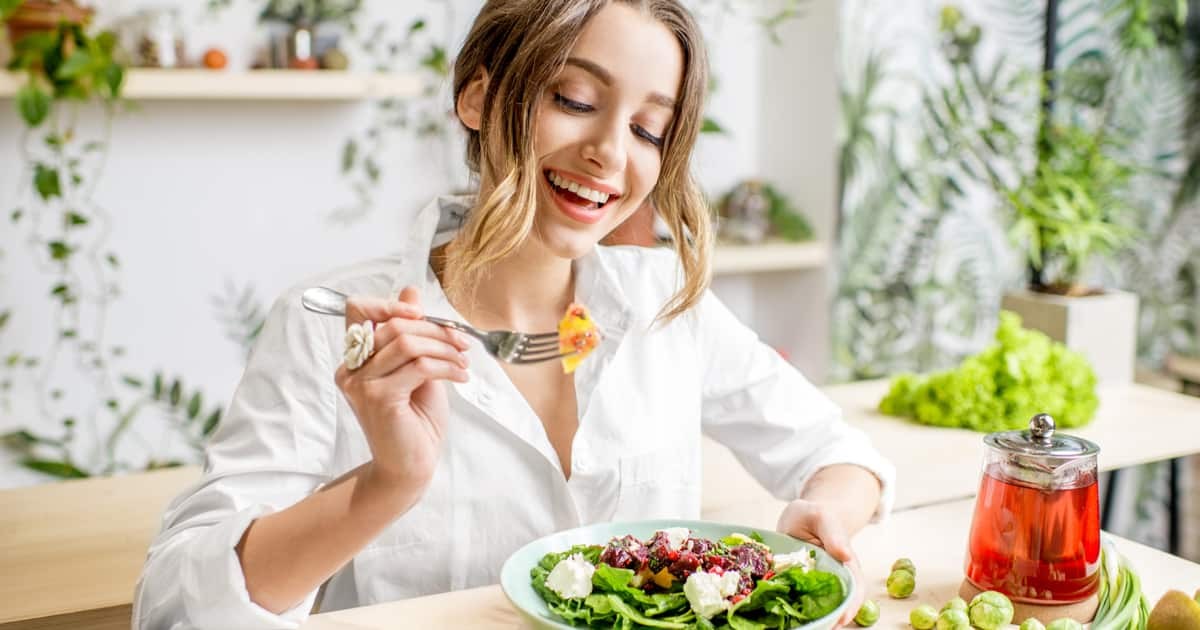 genç bir kadın yeşillik salatası ve bitki çayı bulunan masada otururken gülümseyerek elindeki çatalı ağzına doğru götürüyor.